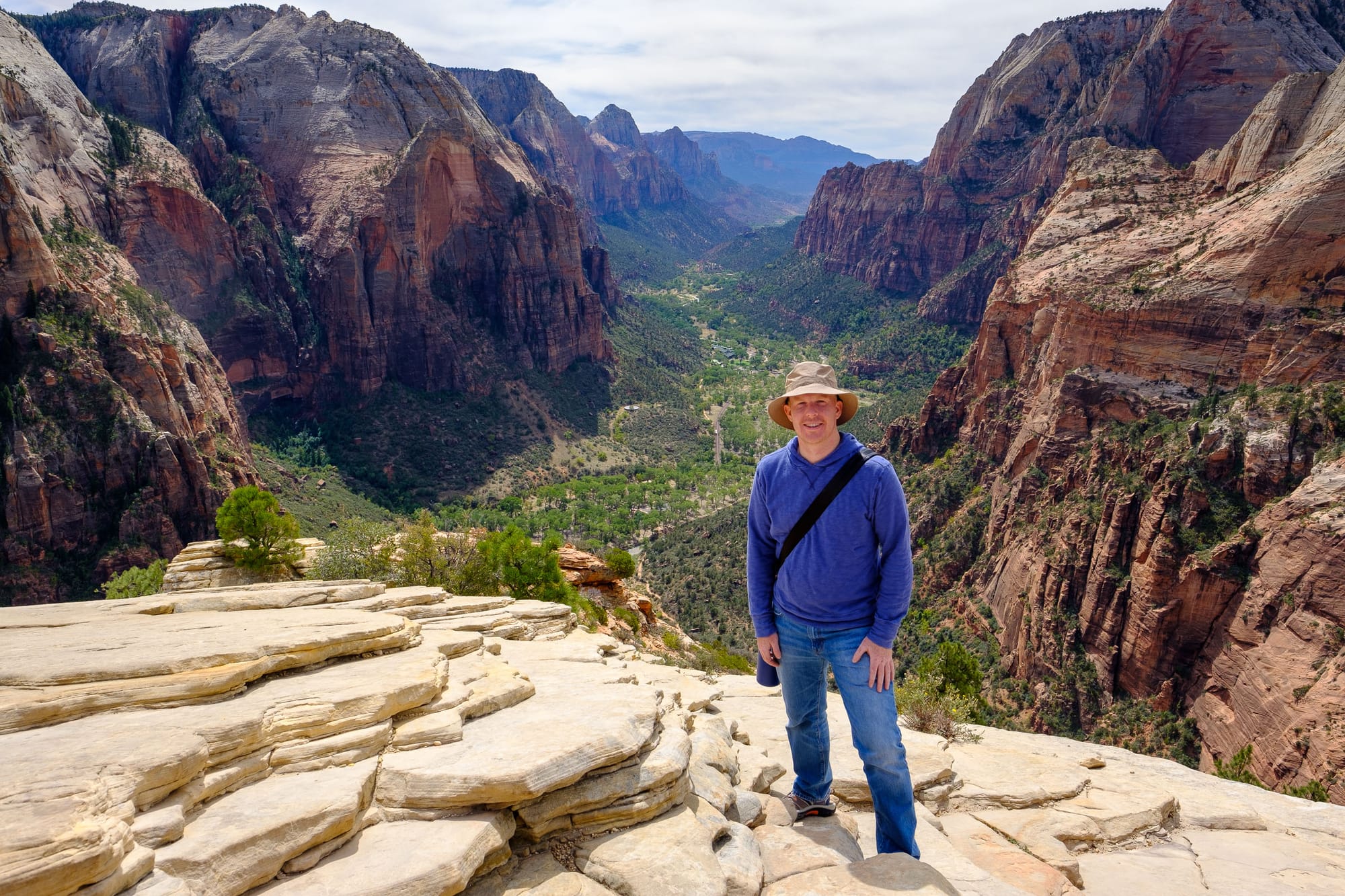 Alex Anderson on top of Angel's Landing in Zion National Park. Red rock cliffs surround a green valley.