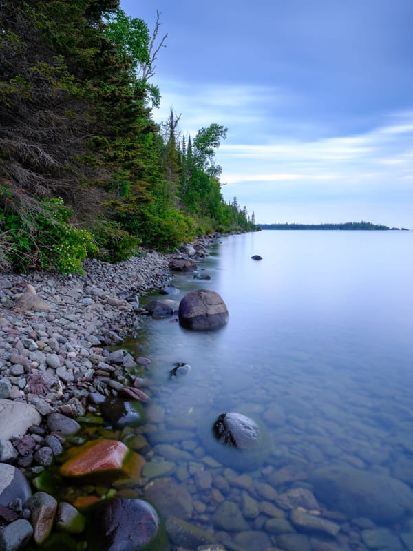 Rock-strewn shoreline at Rock Harbor in Isle Royale National Park, with calm, clear water and dense forest along the coast.