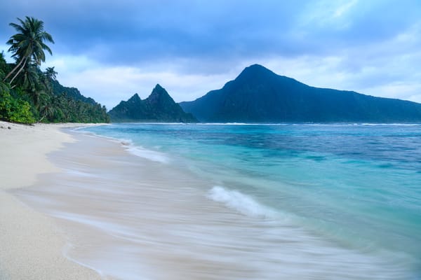 Ofu Beach with soft, cras waves, white sand, palm trees, and lush mountains in the background.